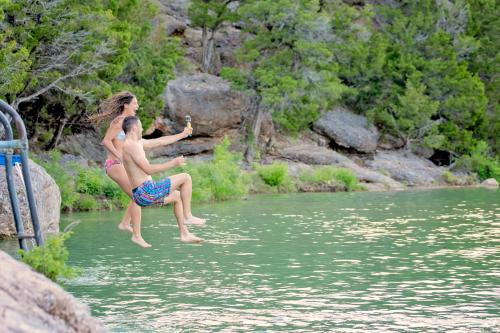 A man and woman jump into a green lake from a rocky shore, surrounded by trees and nature.