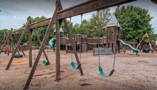 A playground featuring swings, a slide, and wooden structures surrounded by trees and gravel.
