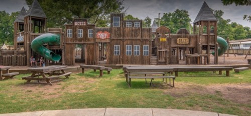 A wooden playground resembling a western town, featuring slides, towers, and picnic tables on a grassy area.