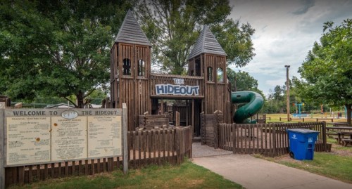 Playground structure called "The Hideout" with wooden towers, a slide, and a welcome sign in a park setting.
