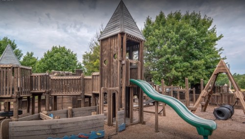 Wooden playground with a slide, climbing structures, and swings, surrounded by trees and gravel ground.