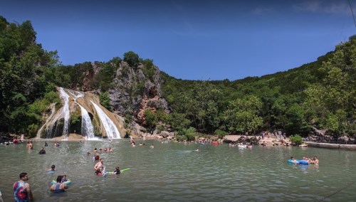 A scenic waterfall surrounded by lush greenery, with people swimming and relaxing in the clear water below.