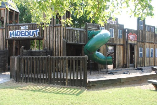 A wooden playground structure with a green slide, featuring signs for "The Hideout" and "Hotel."
