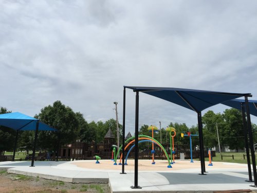 A colorful playground with water features under blue shade structures, surrounded by green trees and a cloudy sky.
