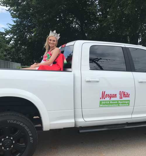 A young woman wearing a crown sits in the back of a white truck, labeled "2019 Rush Springs Watermelon Queen."