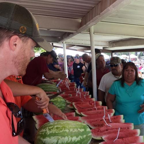 A crowd gathers at a festival, watching as watermelons are sliced and served on a table under a shelter.