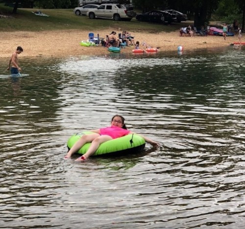 A girl in a pink shirt relaxes on a green float in a lake, surrounded by people enjoying the water and beach.