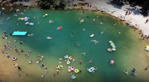 Aerial view of a crowded swimming area with people on floats in a clear green river surrounded by rocks and trees.