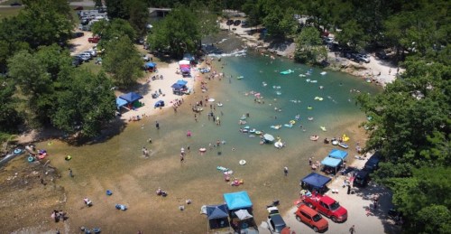 Aerial view of a busy swimming area with people, tents, and colorful floats in a natural setting.