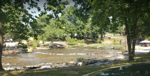A serene river scene with trees, people enjoying the water, and a tent in the background on a sunny day.