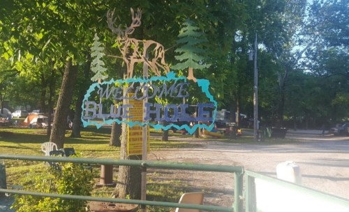 A sign reading "Welcome Blue Hole" with a deer and trees, set in a green outdoor area with camping spots.