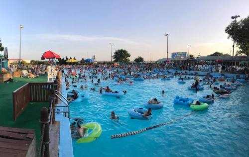 A crowded water park with people floating on inner tubes in a large pool under a clear sky.