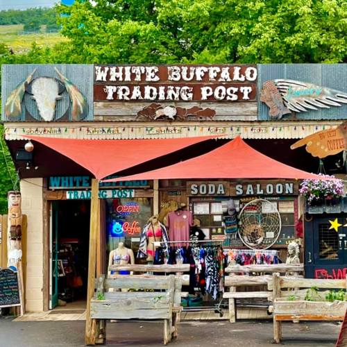 A rustic trading post with a sign reading "White Buffalo Trading Post," featuring outdoor seating and colorful merchandise.
