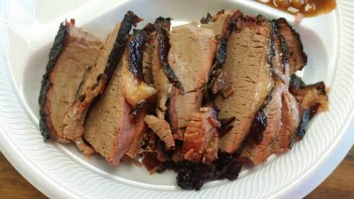 Sliced smoked brisket on a white plate, showcasing tender meat with a smoky crust and a side of barbecue sauce.