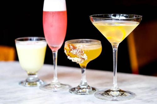Four colorful cocktails on a marble surface: a yellow drink, a pink sparkling drink, a golden cocktail, and a clear martini.