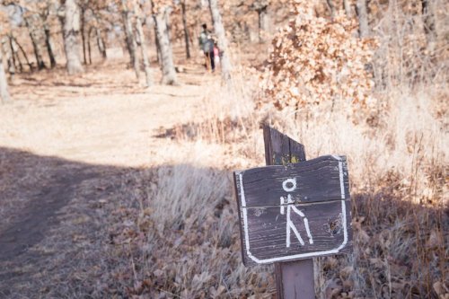 A weathered hiking sign stands in a wooded area with autumn leaves, while two hikers are visible in the background.