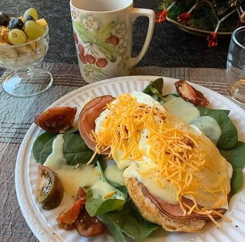 A plate of eggs Benedict on spinach, topped with cheese, alongside a cup of coffee and a fruit dessert.