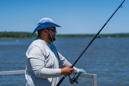 A man in a blue hat and long-sleeve shirt stands by the water, holding a fishing rod and looking out at the lake.