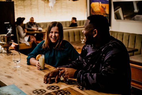 A woman and man laugh together at a bar, enjoying drinks in a cozy, lively atmosphere.