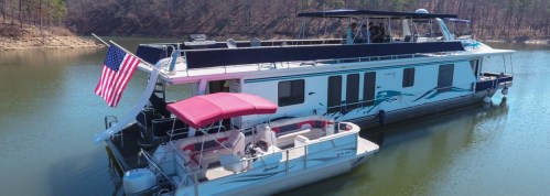 A houseboat with an American flag docked on a calm lake, alongside a smaller pontoon boat. Trees line the shore.