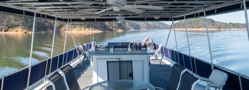 A spacious boat deck with a table and chairs, overlooking calm water and wooded shores in the background.