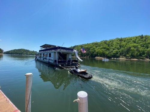 A houseboat navigates a calm lake, surrounded by green hills and a clear blue sky.