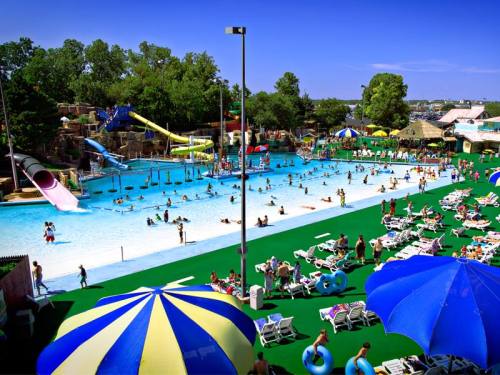 A vibrant water park scene with pools, slides, and sunbathers under a clear blue sky. Colorful umbrellas dot the area.