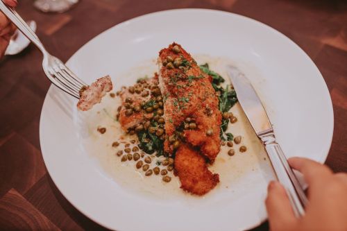 A plate of crispy fried fish on a bed of greens, topped with capers and a creamy sauce, with a fork in hand.