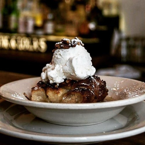 A slice of dessert topped with whipped cream and chocolate sauce, served on a white plate in a cozy setting.