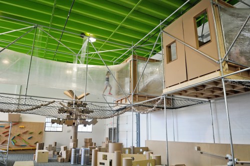 A play area featuring a suspended structure with a net, cardboard playhouse, and a child walking above.