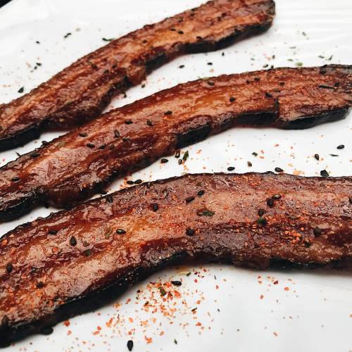 Three crispy strips of glazed meat on a white plate, sprinkled with black sesame seeds and seasoning.