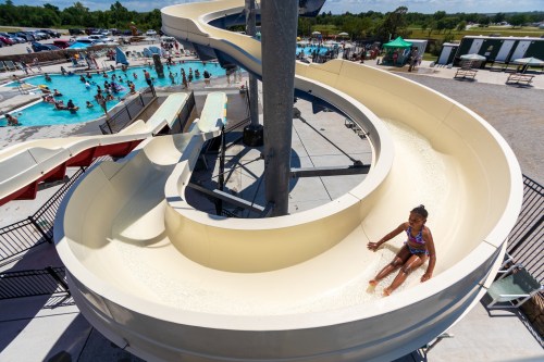 A child slides down a water slide at a busy pool area on a sunny day, with people enjoying the water below.