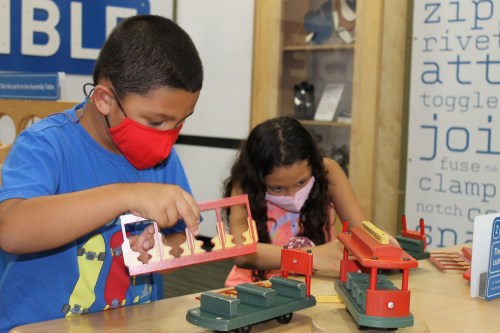 Two children play with wooden building toys, focused on assembling structures, wearing masks in a bright indoor space.