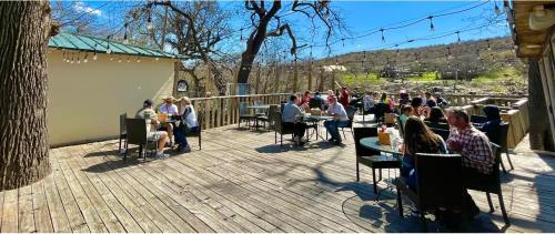 Outdoor dining area with people seated at tables on a wooden deck, surrounded by trees and a clear blue sky.