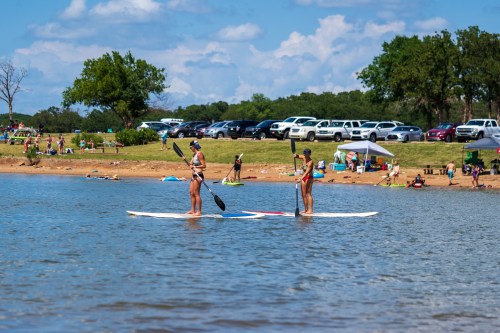 Two women paddleboard on a calm lake, with a beach and parked cars in the background under a blue sky.