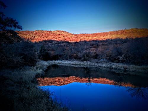 A serene landscape featuring a calm pond reflecting autumn-colored hills under a clear blue sky.