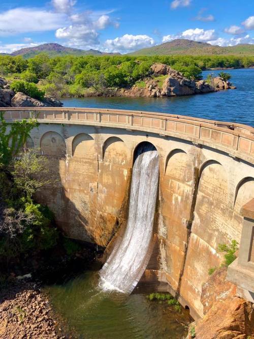 A dam with a waterfall cascading over its edge, surrounded by lush greenery and a serene blue lake under a clear sky.