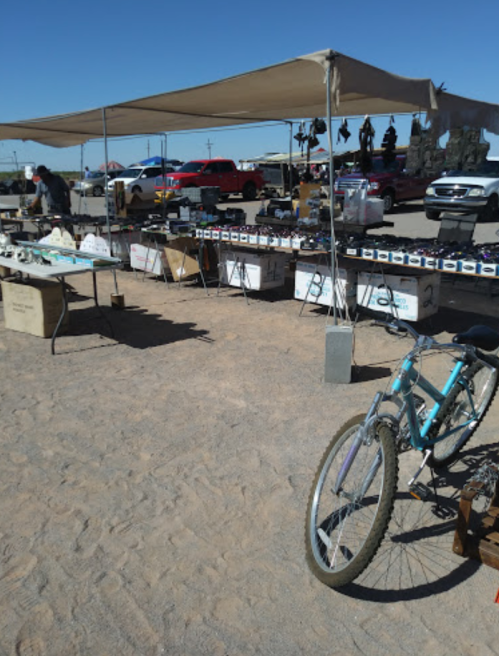 A sunny outdoor market with tables of items for sale, a bicycle in the foreground, and vehicles in the background.