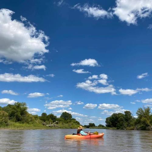 A person kayaking on a calm river under a bright blue sky with fluffy white clouds and green trees in the background.