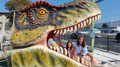 A man and two girls pose inside a colorful dinosaur sculpture, smiling in bright sunlight.