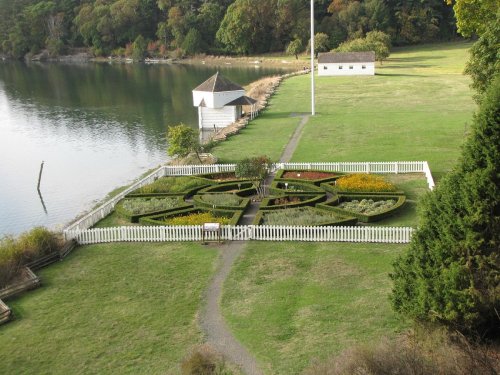 Aerial view of a landscaped garden with a circular design, surrounded by a white picket fence, near a calm water body.