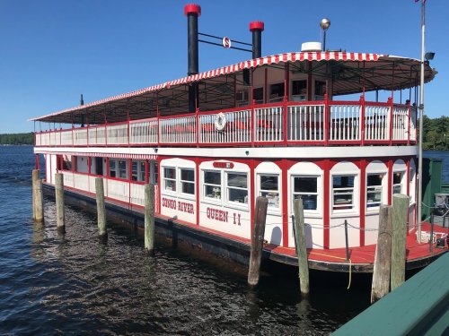 A red and white riverboat docked on calm water, featuring a striped awning and multiple decks.