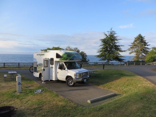 A parked RV by the ocean, surrounded by trees and a clear sky, with a picnic area nearby.