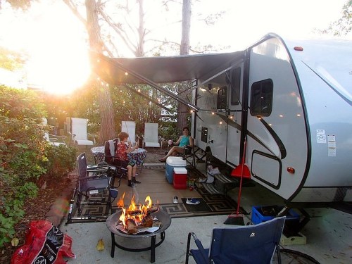 A cozy campsite with a trailer, outdoor seating, and a fire pit surrounded by trees and soft lighting.