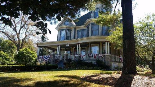 A large Victorian-style house with a wraparound porch, surrounded by greenery and decorated with red, white, and blue bunting.