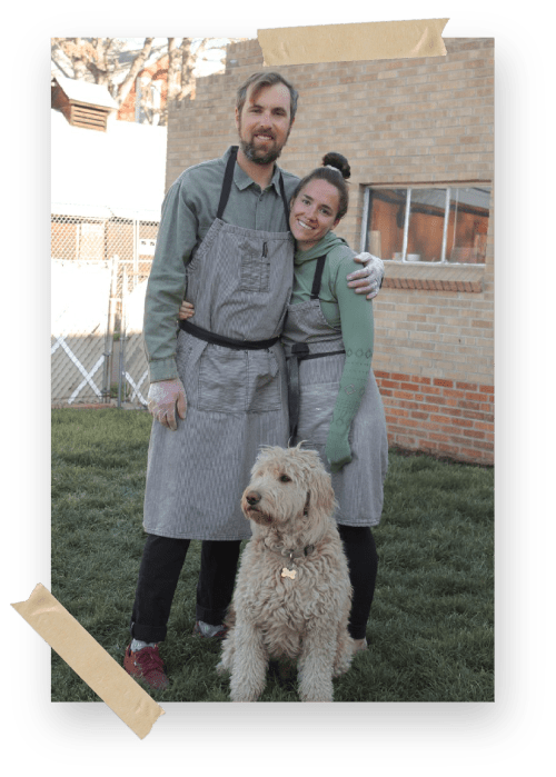 A couple in matching aprons stands together with a dog on grass, smiling at the camera.
