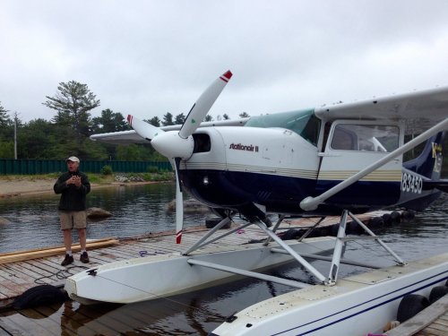 A man stands beside a blue and white seaplane on a dock by a calm lake, surrounded by trees and overcast skies.