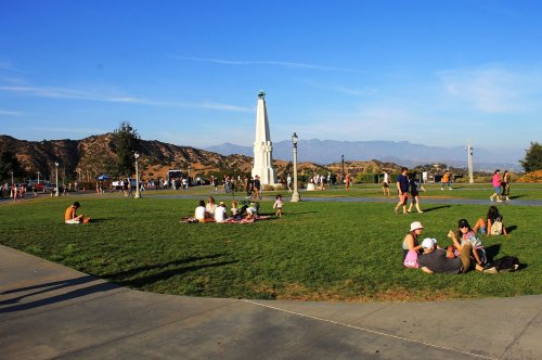 People relax on a grassy park area with a monument, surrounded by mountains and a clear blue sky.