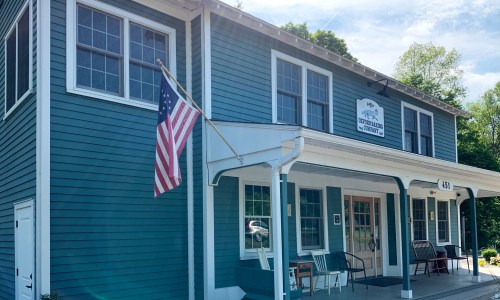 A blue building with white trim, featuring an American flag and a sign, surrounded by greenery.