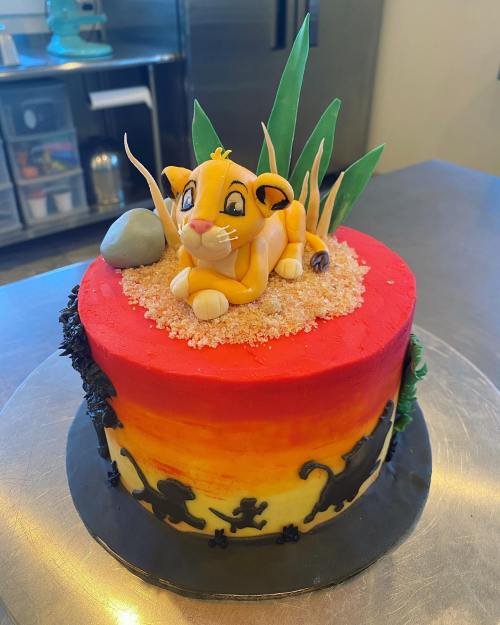 A colorful cake featuring a fondant lion cub on top, surrounded by grass and decorative elements.